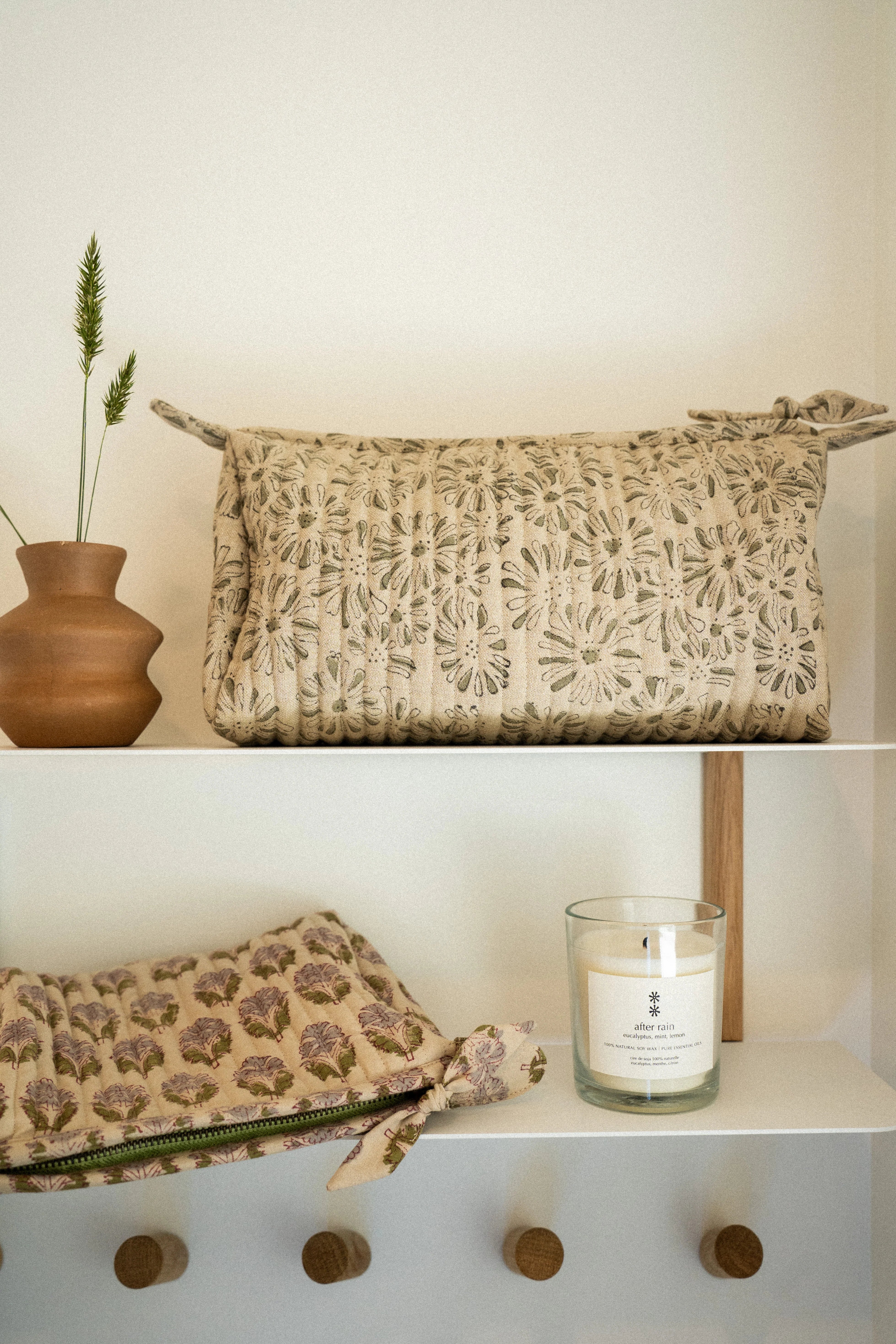 Woven clutch bag and candle on a white shelf with wooden hooks.